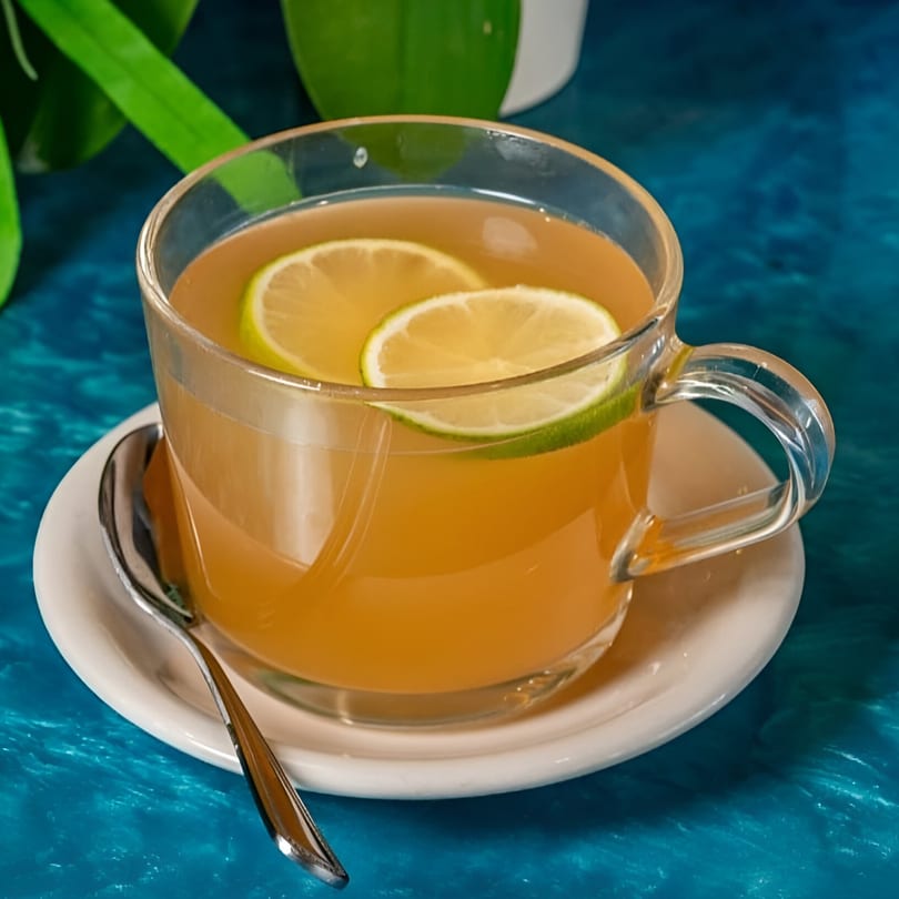 A clear mug of hot tea with two lime slices, on a white saucer with a spoon.