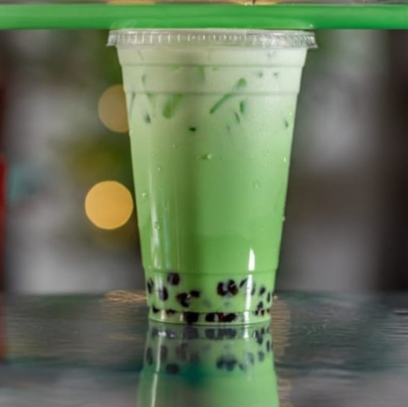 Green boba tea with black tapioca pearls in a clear plastic cup on a reflective surface.
