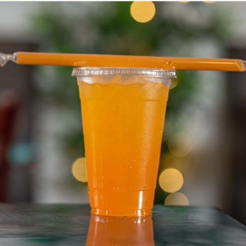A vibrant orange drink in a clear plastic cup with a straw, reflecting on a dark surface.